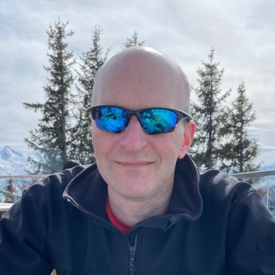 Photograph of James Bull, taken outside with a background trees, mountains and a cloudy sky. He is smiling at the camera and wearing blue tinted glasses.